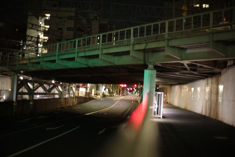 Road under a green rail bridge with motion blur and empty sidewalks at night